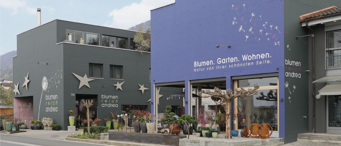 A purple building with the words 'Blumen. Garten. Wohnen.' on it. Next to it is a gray building with white stars. The purple building has potted plants and a tree in front of it.