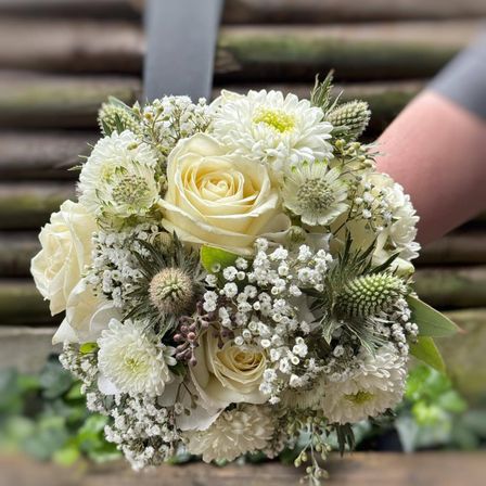 Ein Nahaufnahme eines Brautstraußes mit weißen Rosen, Gänseblümchen, Babysatem und Disteln mit einem unscharfen hölzernen Hintergrund.