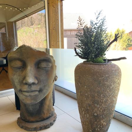 In einem Raum mit großen Fenstern steht eine steinerne Kopfskulptur mit geschlossenen Augen neben einem großen Terrakotta-Gefäß, das eine grüne Pflanze mit einem Zweig enthält.