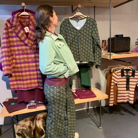 Eine Frau steht in einem Bekleidungsgeschäft und betrachtet einen bunten Mantel und ein gemustertes Oberteil an Kleiderbügeln, während verschiedene Kleidungsstücke und Accessoires um sie herum ausgestellt sind.