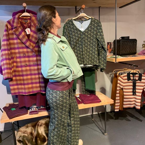 A woman stands in a clothing store, gazing at a colorful coat and a patterned blouse on hangers, while various clothes and accessories are displayed around her.