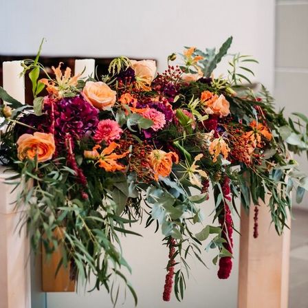 Eine Blumendekoration auf einem Tisch besteht aus orangen, roten und lilafarbenen Blumen mit grünen Blättern, akzentuiert durch zwei hohe weiße Kerzen.