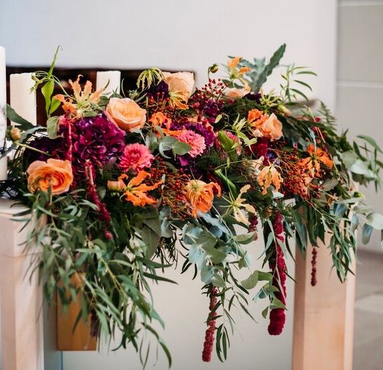 A floral arrangement on a table includes orange, red, and purple flowers with green leaves, accented by two tall white candles.