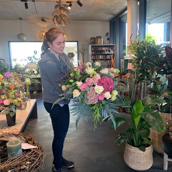 A woman is holding a large bouquet of flowers in a flower shop. Various potted plants and flowers are around her.