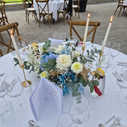 Ein Tisch ist mit einem Blumenarrangement aus blauen und weißen Blumen, weißen Rosen und grünen Blättern dekoriert. Es gibt Weinläser, eine Zitronenscheibe und zwei brennende Kerzen. Vor dem Arrangement steht ein Menü. Im Hintergrund sind weitere Tische gedeckt.