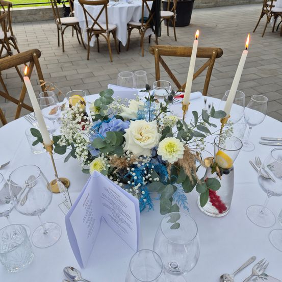 Ein Tisch ist mit einem Blumenarrangement aus blauen und weißen Blumen, weißen Rosen und grünen Blättern dekoriert. Es gibt Weinläser, eine Zitronenscheibe und zwei brennende Kerzen. Vor dem Arrangement steht ein Menü. Im Hintergrund sind weitere Tische gedeckt.