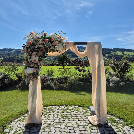 Ein mit Blumen geschmückter Hochzeitsbogen steht auf einem Kiesweg in einem üppigen grünen Feld unter einem blauen Himmel mit fernen Hügeln.