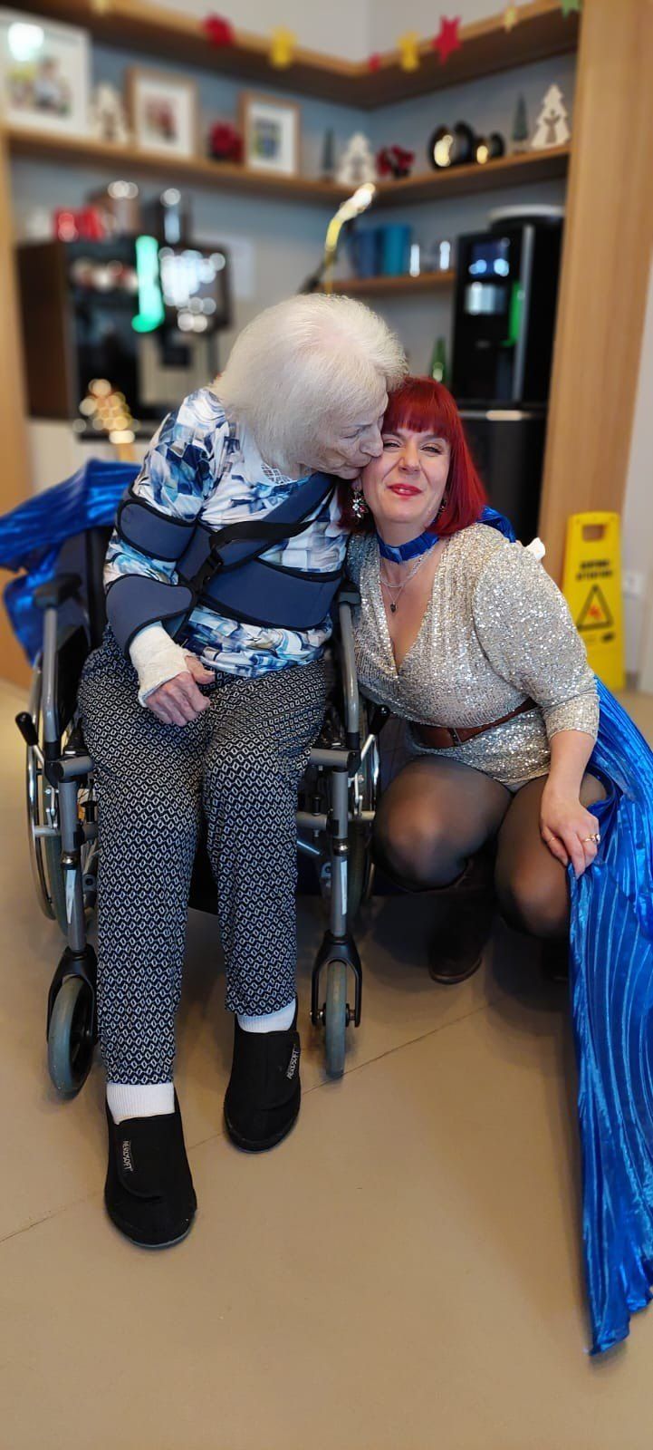 An elderly woman in a wheelchair is being hugged by a woman in a sequined dress. Both are smiling. Behind them is a yellow sign with a warning symbol.