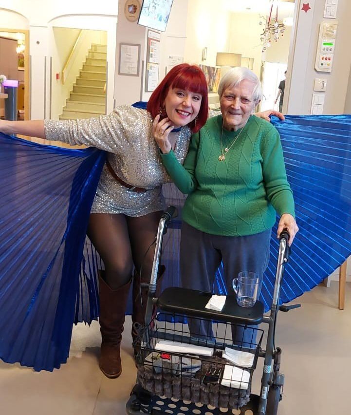 Two women pose for a photo. One in a glittery costume with wings, and the other in a green sweater, both smiling. They stand near a wheeled cart with a cup and papers.