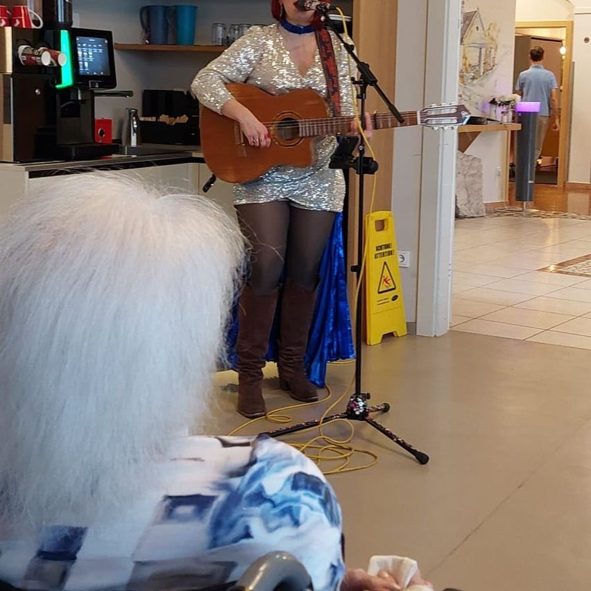 A woman is playing a guitar on stage in front of a seated audience. Behind her is a kitchen area with a coffee machine and a shelf.