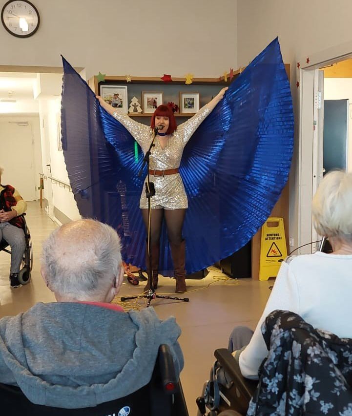 A woman with red hair in a silver outfit performs on stage with large blue wings. She stands in front of a microphone with a stand. Behind her, there is a shelf with framed pictures. Several elderly people sit in wheelchairs and watch her.