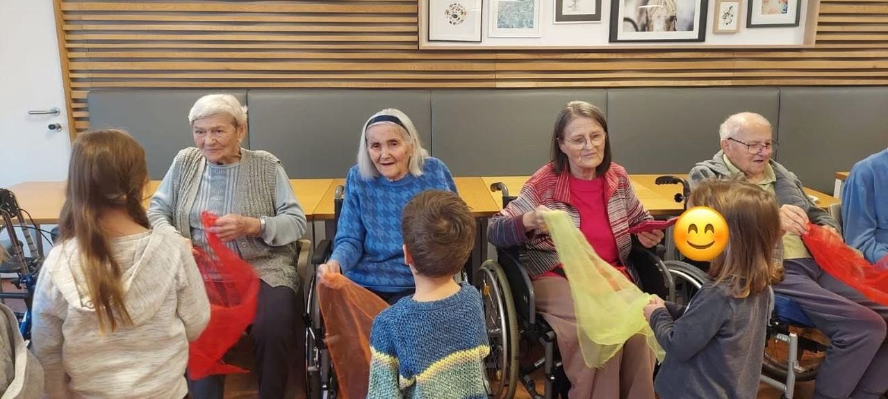 Drei ältere Frauen sitzen in Rollstühlen, eine hält ein rotes Tuch, eine andere ein gelbes, während ein Kind zwischen ihnen steht. Sie befinden sich in einem Raum mit Holzwänden und gerahmten Bildern.