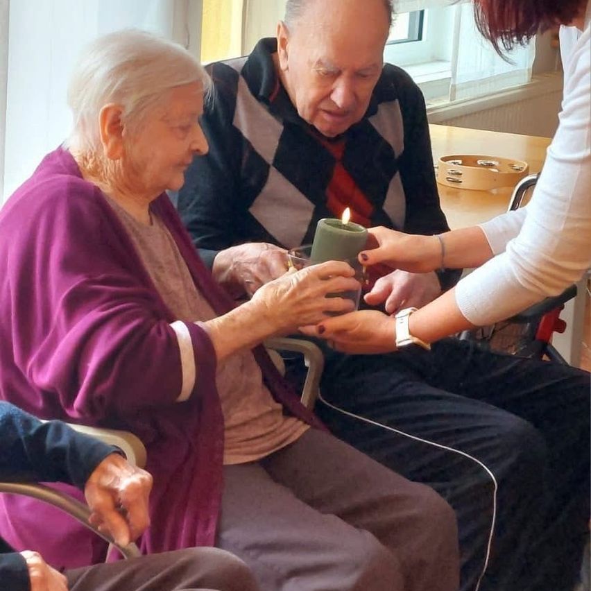 Zwei ältere Menschen sitzen zusammen und halten eine Kerze, eine Frau lächelt in die Kamera. Eine ältere Frau hält eine Kerze mit einer Helferin neben ihr. Sie sind in einem Raum mit einem Tisch und Fenster im Hintergrund.