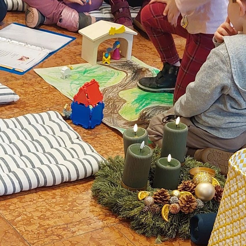 Mehrere Kinder sitzen auf dem Boden, vor ihnen liegt ein Kranz aus grünen Blättern, Kerzen und Ornamenten. Auf dem Boden befindet sich auch eine Zeichnung.