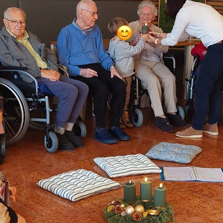 Ältere Menschen sitzen mit einem kleinen Kind in Rollstühlen und lächeln. Eine Frau hält eine Kerze und ein kleines lächelndes Gesicht in der Hand. Kerzen und eine Girlande liegen auf dem Boden.