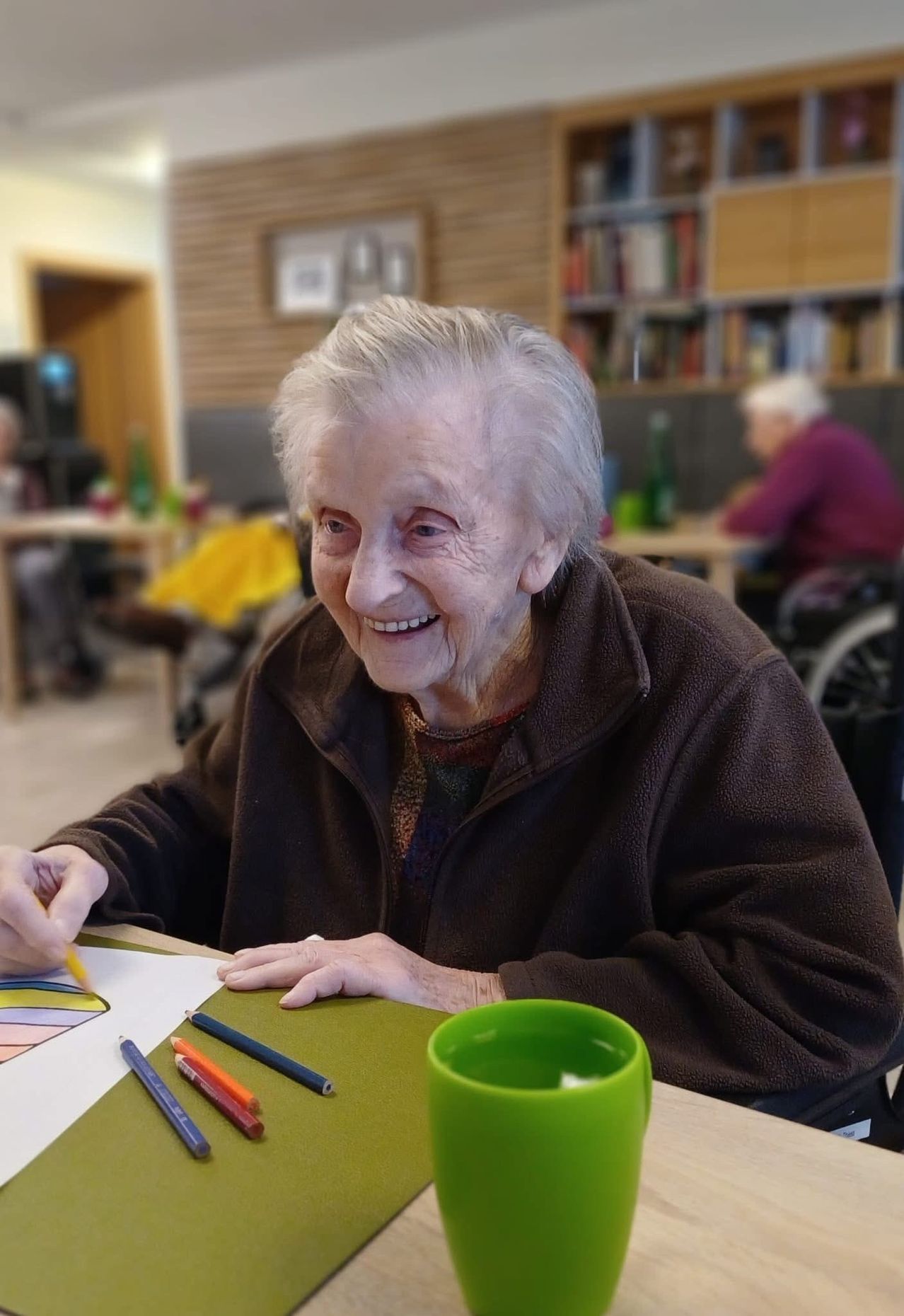 An elderly woman smiles while seated in a wheelchair, drawing on a table with colored pencils and a green cup nearby.