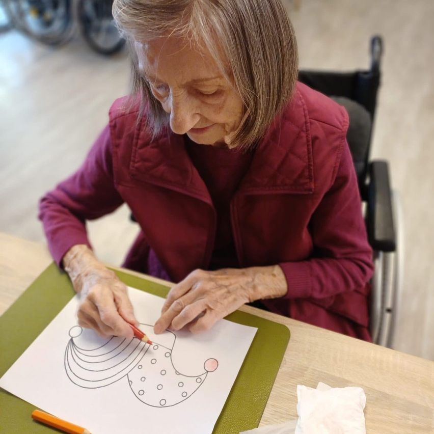 An elderly woman in a wheelchair colors a whimsical drawing on a paper placed on a green mat on a table.