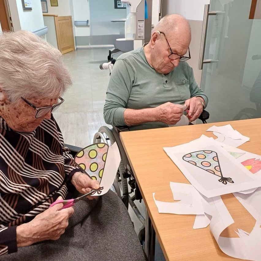Two elderly individuals are crafting party hats, seated in wheelchairs. One uses scissors while the other holds a hat. Various colored paper pieces are on the table. The room has white walls and tile flooring.