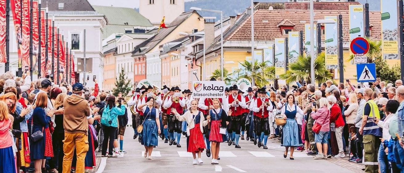 Ein festlicher Umzug findet in einer europäischen Stadt mit einem Kirchturm im Hintergrund statt. Viele Menschen stehen auf beiden Seiten der Straße und beobachten den Umzug. Der Umzug besteht aus Menschen in traditionellen Kostümen, die Musikinstrumente spielen.