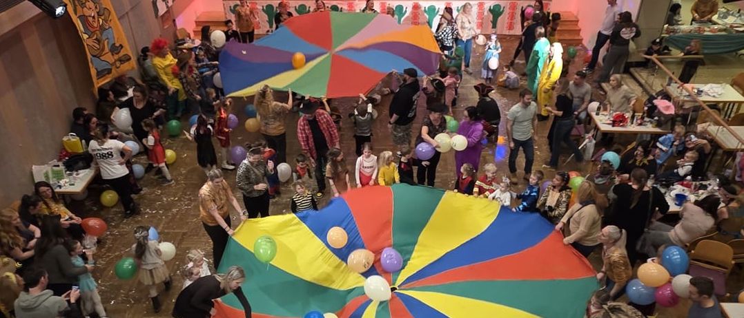 Ein Blick von oben auf eine Menschenmenge, die in einem Theater mit Bühne, farbenfrohen Schirmen und Ballons versammelt ist. Die Bühne hat eine Kulisse und ein Tipi.
