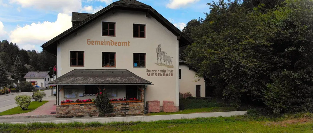 Ein Gemeindeamt mit einer Wandmalerei eines Mannes und einer Ziege, umgeben von Grün und Bäumen.