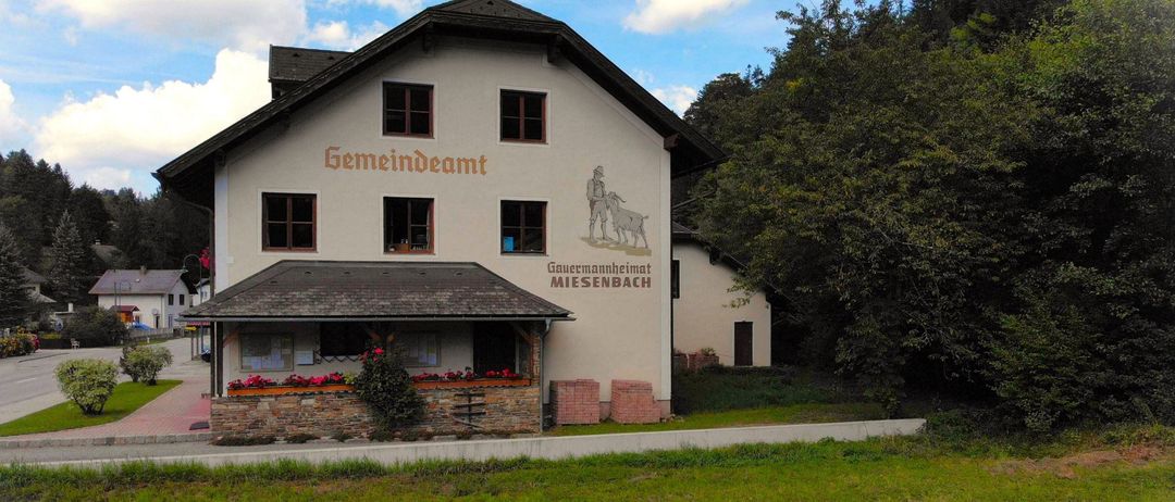 Ein Gemeindeamt mit einer Wandmalerei eines Mannes und einer Ziege, umgeben von Grün und Bäumen.