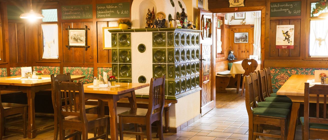 Ein gemütliches Restaurant-Interieur mit Holzmöbeln, gefliesten Böden und dekorativen Gegenständen. Der Tisch ist gedeckt, und es sind Stühle aufgestellt. An der Wand befindet sich ein Menübrett, und ein Schrank hat grüne Glasfächer.