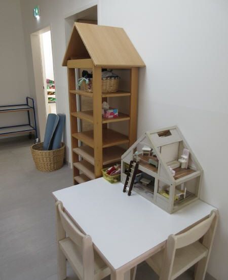 Ein Raum mit einem weißen Tisch und Stühlen, einem Puppenhaus auf dem Tisch, einem Holzregal, einem Korb und einer Yogamatte.