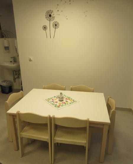 Ein Esszimmer mit einem quadratischen weißen Tisch und sechs Stühlen. Ein dekoratives Serviettentuch und eine Keramikvase liegen auf dem Tisch. Der Raum hat eine weiße Wand mit Löwenzahn-Aufklebern. Ein Waschbecken befindet sich auf der linken Seite.