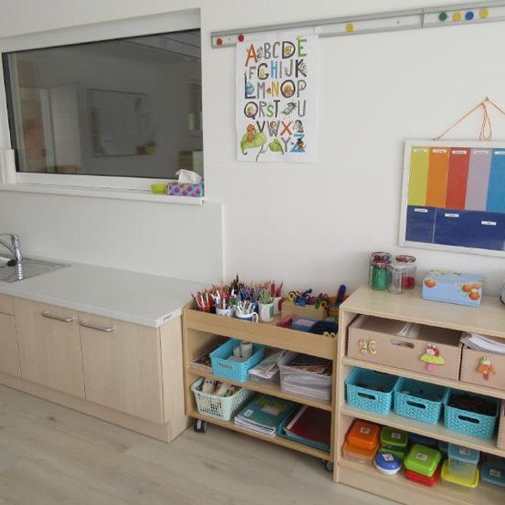 Eine Küchenecke mit einem Waschbecken, Schränken und Regalen mit Spielzeug und Papieren. An der Wand hängt ein buntes Alphabet-Poster.