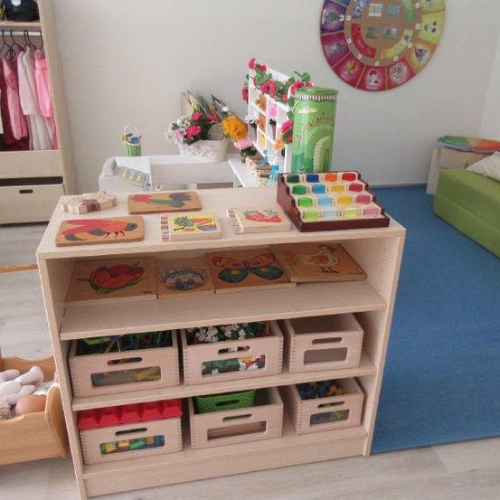 Ein Raum mit einem Holzregal voller Spielzeug und Aufbewahrungsboxen, einem blauen Teppich, einer grünen Couch mit Kissen, einem grünen Stuhl, einem Kleiderschrank mit Kleidung und einem runden dekorativen Stück an der Wand.