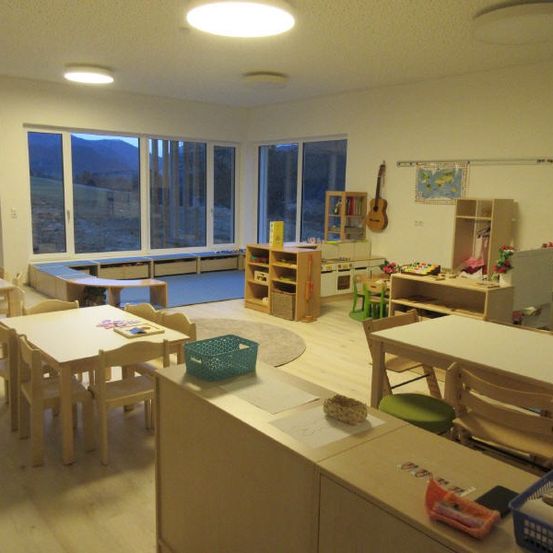 Ein Klassenzimmer mit Holztischen und Stühlen, großen Fenstern mit Blick auf die Berge und verschiedenen Lernspielzeug und einer Gitarre an der Wand.