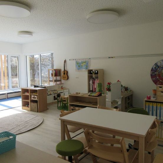 Ein heller Raum mit weißen Wänden, Holzmöbeln und einem Teppich. Es gibt einen Tisch, Stühle, eine Gitarre, Regale mit Büchern und Spielzeug und ein großes Fenster mit Blick auf Bäume draußen.