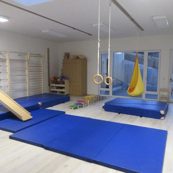 Ein Gymnastikraum mit blauen Matten, einer Holzrutsche, hängenden Ringen und einem gelben Schaukel. Holzregale und Aufbewahrungsboxen stehen in der Nähe der Glastüren.