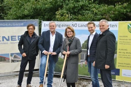 Fünf Personen posieren mit Schaufeln für ein Foto auf einer Baustelle mit einem Banner dahinter.