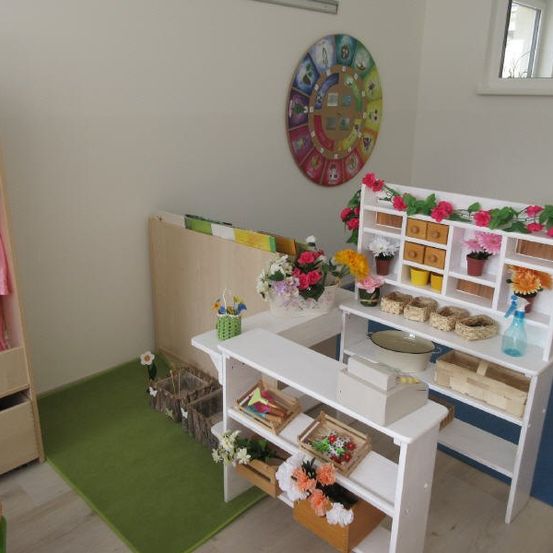 Ein Raum mit einem grünen Teppich, einem weißen Regal mit Blumen, einer runden Uhr, einem Holzschrank mit Kleidern und einem Spiegel an der Wand.