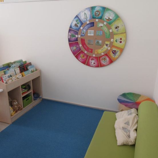 Ein Kinderspielzimmer mit blauem Teppich, einem Bücherregal voller Bücher und einer kreisförmigen Wanddekoration. Ein grünes Sofa mit Kissen und ein bunter Hocker befinden sich rechts.
