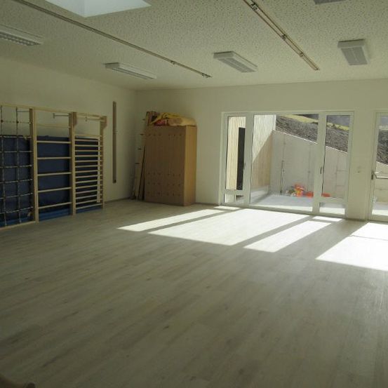 Ein leerer Raum mit Holzboden, großen Fenstern und Fitnessgeräten auf der linken Seite. Es gibt einen Holzschrank an der Wand und Lampen an der Decke.