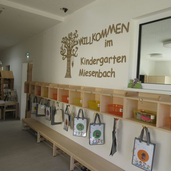 Ein Kindergartenraum mit einem Baum und Text an der Wand. Taschen und Körbe hängen an der Wand. Es gibt eine Bank und Regale. Ein Spiegel reflektiert einen Raum.