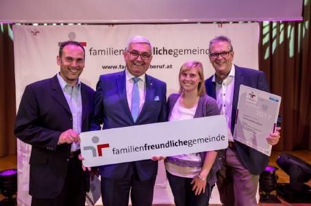 Vier Personen in Anzügen und Krawatten stehen auf einer Bühne und halten ein Zertifikat und ein großes weißes Banner mit Text. Das Banner trägt den Schriftzug 'familienfreundlichegemeinde' und ein Logo. Dahinter befindet sich eine Kulisse mit einer Website-Adresse.