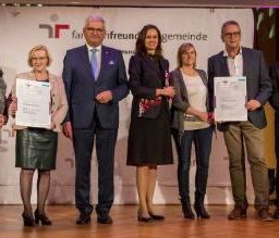 Eine Gruppe von Menschen in formeller Kleidung steht auf der Bühne, hält Urkunden in den Händen, mit einem Banner hinter ihnen.