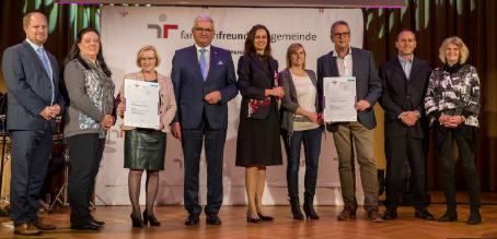 Eine Gruppe von Menschen in formeller Kleidung steht auf der Bühne, hält Urkunden in den Händen, mit einem Banner hinter ihnen.