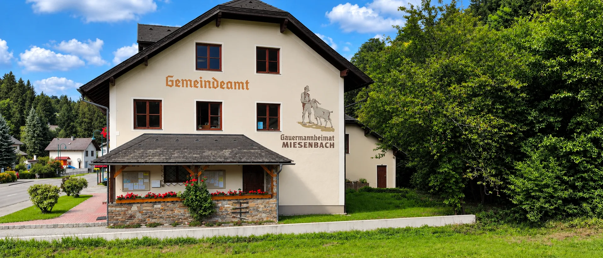 Ein weißes Gebäude mit schwarzem Dach und der Aufschrift 'Gemeindeamt'. Vor dem Gebäude befindet sich ein kleiner Vorbau mit Blumen.