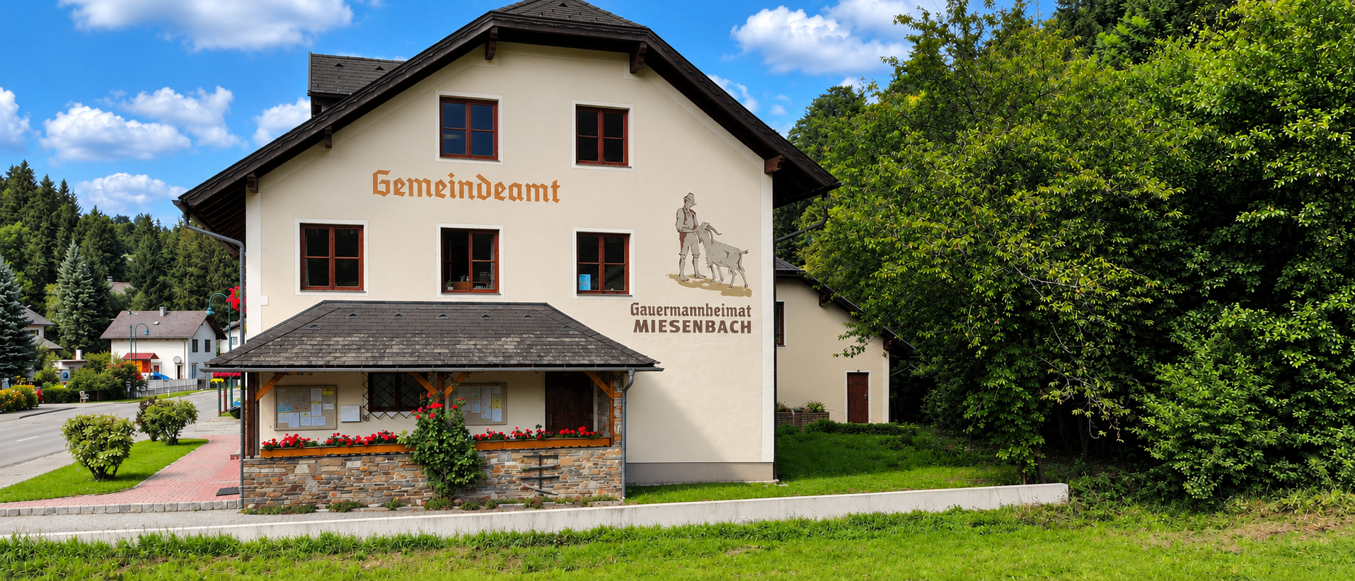 Ein weißes Gebäude mit schwarzem Dach und der Aufschrift 'Gemeindeamt'. Vor dem Gebäude befindet sich ein kleiner Vorbau mit Blumen.