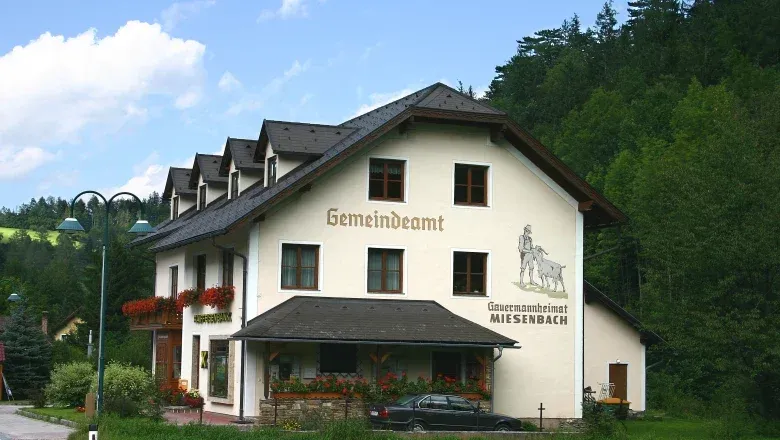 Ein weißes Gebäude mit schwarzem Dach, Gemaindeamt auf der Wand, Blumen davor und ein schwarzes Auto davor.