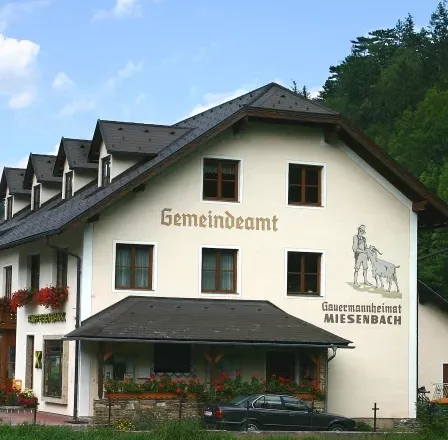 Ein weißes Gebäude mit schwarzem Dach, Gemaindeamt auf der Wand, Blumen davor und ein schwarzes Auto davor.