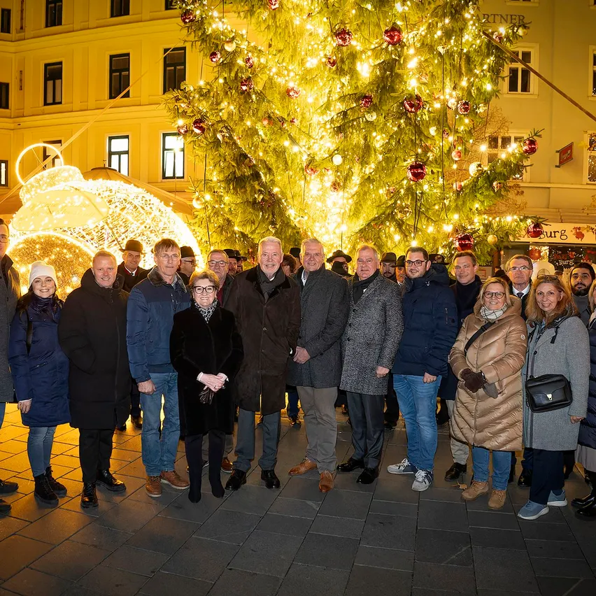 Eine Gruppe von Menschen in Winterkleidung steht zusammen vor einem großen Weihnachtsbaum, der mit Lichtern und Ornamenten beleuchtet ist. Dahinter befindet sich ein Gebäude mit vielen Fenstern.