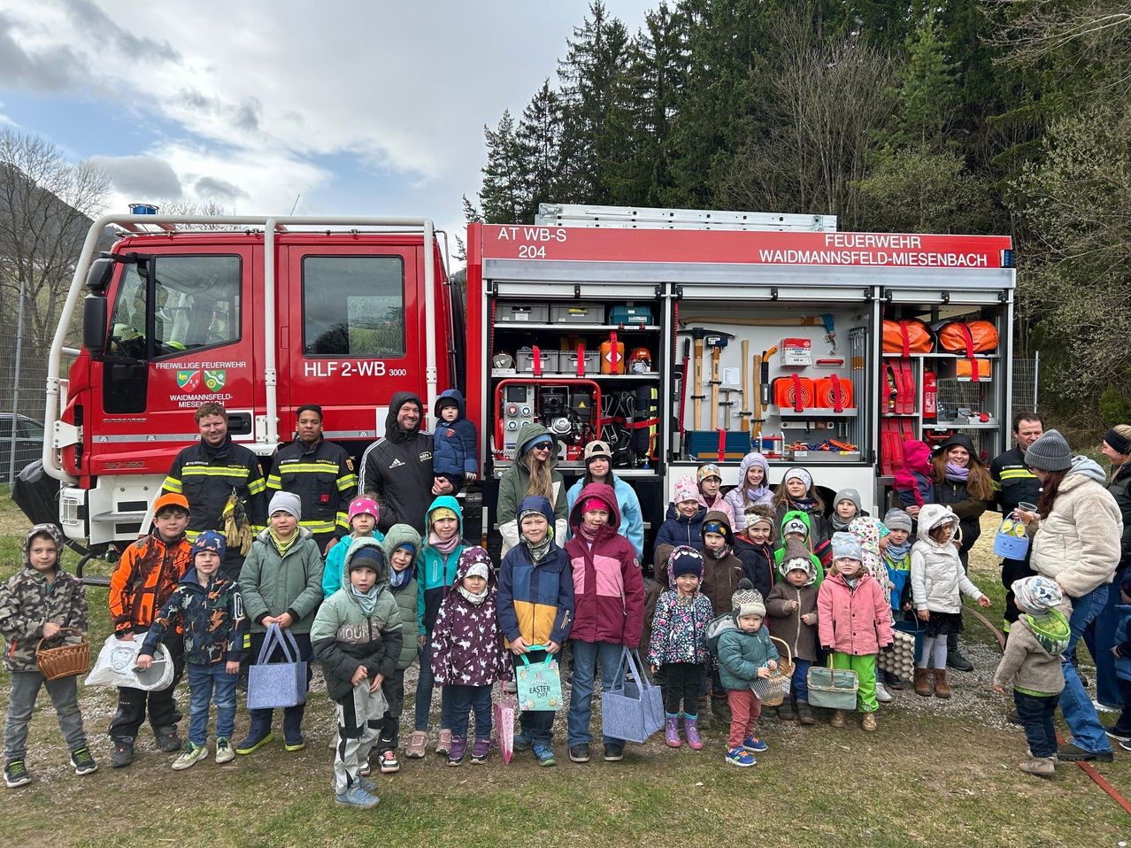 Eine Gruppe von Kindern und Erwachsenen posiert mit einem Feuerwehrwagen, hält Osterkörbchen und trägt Winterkleidung. Bäume und ein bewölkter Himmel im Hintergrund.