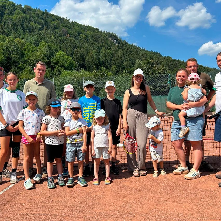 Eine Gruppe von Menschen, einschließlich Kindern, posiert für ein Foto auf einem Tennisplatz mit einem Netz im Hintergrund. Sie sind in Freizeitkleidung gekleidet, und einige halten Tennisschläger.