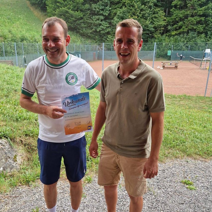 Zwei Männer stehen auf einem Kiesweg, einer hält ein Zertifikat in der Hand, beide lächeln. Hinter ihnen befindet sich ein Tennisplatz mit einem Zaun und viel Grün.